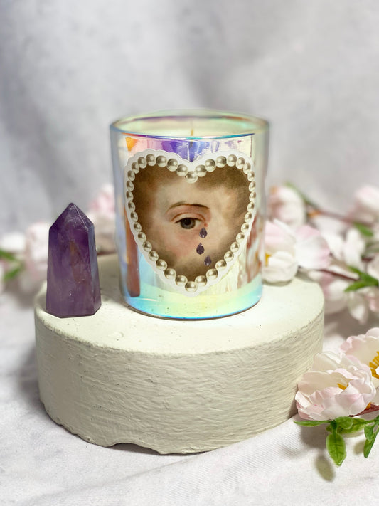 Candle with a heart-shaped cutout of a eye, surrounded by flowers and an amethyst crystal on a textured surface.